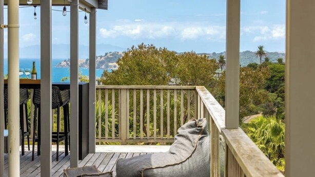 Una terraza de madera con un sillón de mimbre grisáceo y una pequeña mesa con sillas, con vistas a un paisaje panorámico del océano y las colinas.