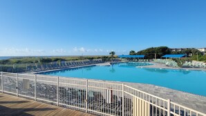 En stor udendørs swimmingpool med en træterrasse og hvidt hegn, omgivet af liggestole og parasoller.