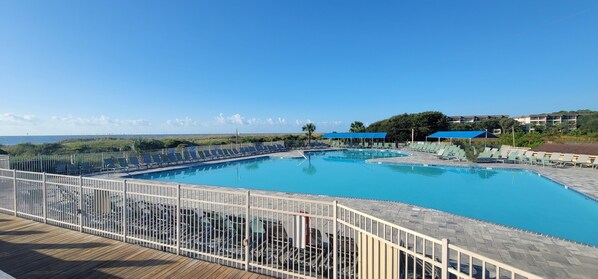 A large outdoor swimming pool with a wooden deck and white fencing, surrounded by lounge chairs and umbrellas.