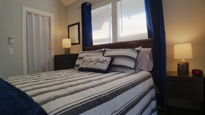 Cama bem arrumada com roupa de cama listrada, mesa de cabeceira com abajur e janela com cortinas.