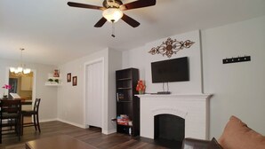 Uma sala de estar com lareira, ventilador de teto, televisão montada acima e um espaço de jantar com cadeiras e mesa.