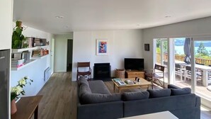 En moderne stue med en grå sofa, et sofabord og et fladskærms-TV. Der er en pejs, en bogreol og et spiseområde med udendørs siddepladser, der kan ses gennem store vinduer.
