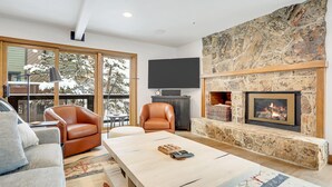 Een moderne woonkamer met een stenen open haard, een flatscreen televisie erboven, leren fauteuils en een groot raam met uitzicht op een terras.