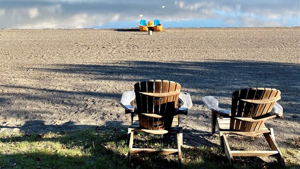 A lakeside view with two wooden chairs facing the water, a row of yellow and blue paddle boats in the distance, and a calm sky.