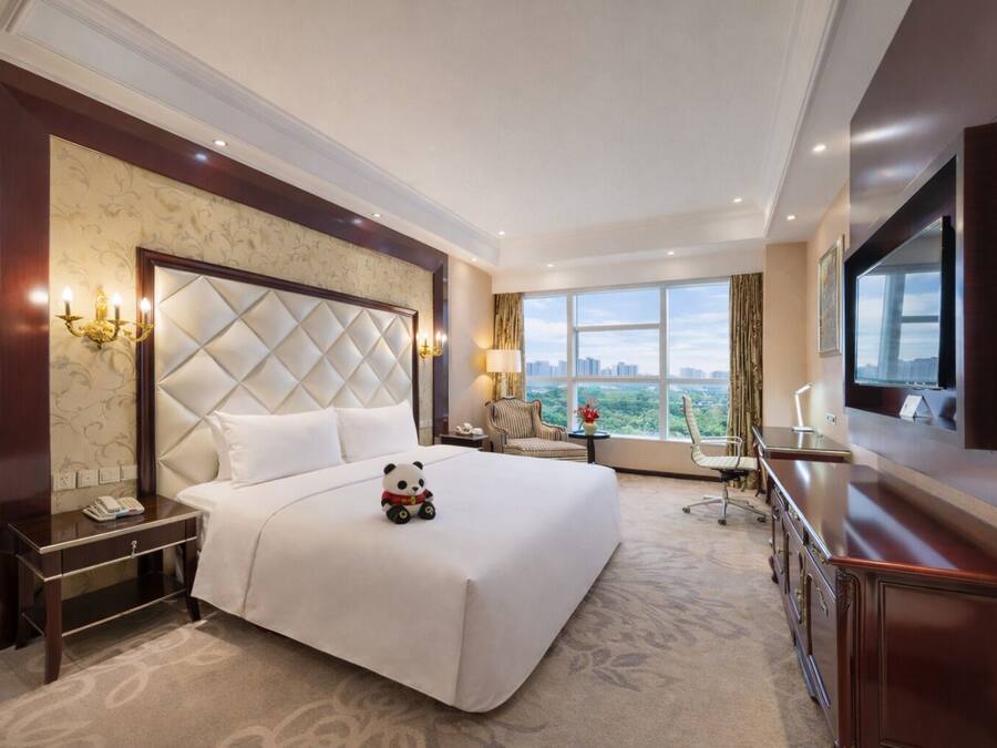 A hotel room with a large bed, a TV, a desk, and a view of the city.