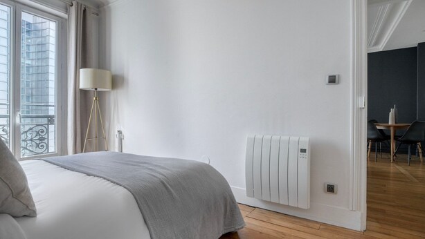 Une chambre à coucher moderne avec un lit, une lampe de chevet, un radiateur et une vue sur un balcon doté d’une balustrade.