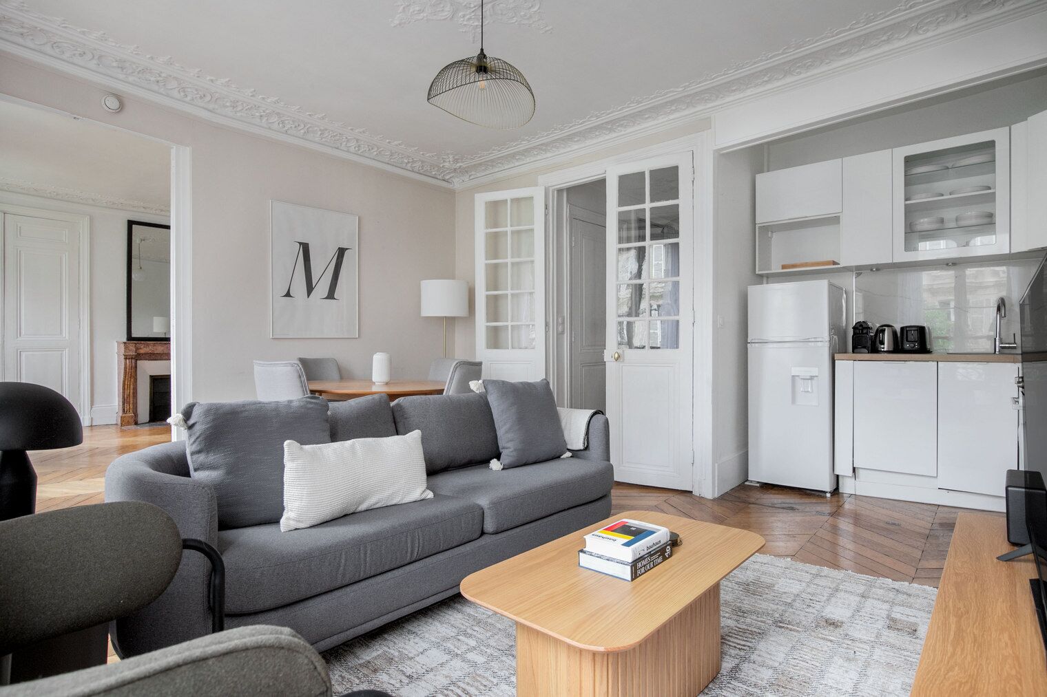 A modern living room with a grey sofa, a wooden coffee table, and a kitchen area with white cabinets and a refrigerator.