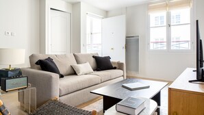 Un salon moderne avec un canapé, une table basse et une petite table sur laquelle se trouve un livre. On y trouve également une lampe, un haut-parleur et un téléviseur posé sur un meuble.