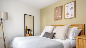 Un lit bien fait, recouvert d’une couverture grise, une table de chevet avec une lampe, et deux œuvres d’art abstraites encadrées accrochées au mur.