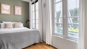 Un dormitorio con una cama grande, dos cuadros en la pared, una mesita de noche con lámpara y una ventana con cortinas.
