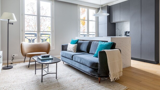 A modern living room with a sofa, coffee table, and a lamp. There is a kitchen area with cabinets and a hanging light fixture. - Blueground | Invalides, view of Matignon Gardens (Paris)
