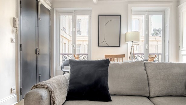 A modern living room with a beige sofa, a black pillow, and a minimalist painting on the wall. - Blueground | Marais, next to Rivoli St shopping (Paris)