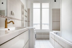 A modern bathroom with a white sink, gold faucet, and a large window.
