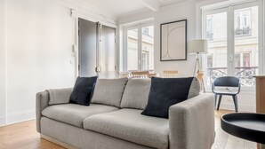 A modern living room with a grey sofa, two black cushions, a wooden chair, and a small black table. - Blueground | Marais, next to Rivoli St shopping (Paris)