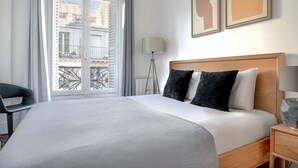 A hotel room with a large bed, a wooden headboard, a bedside lamp, and a view of the city through a balcony door. - Blueground | Marais, next to Rivoli St shopping (Paris)