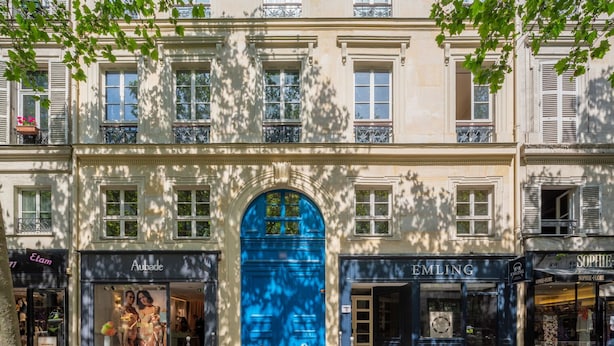 Un immeuble avec une porte bleue bien visible, des devantures de magasins « Etam » et « Emling », et un balcon apparent doté d’une balustrade.