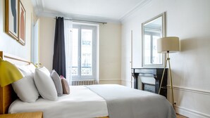 Ein Hotelzimmer mit Bett, Nachttisch, Lampe und Blick auf den Eiffelturm durch das Fenster.