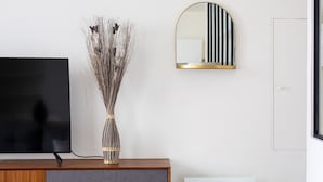 Un salon moderne avec un téléviseur à écran plat posé sur un meuble en bois, un vase décoratif contenant des plantes séchées et un miroir encadré d’or accroché au mur.