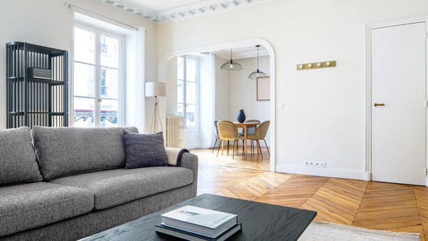 Un salon moderne avec un canapé gris, une table basse avec des livres, un coin repas avec une table et des chaises, et une porte blanche.