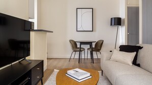 A modern living room with a flat-screen TV, a sofa, a coffee table with a book, and a dining area with chairs. - Blueground | Madeleine, elev, near art (Paris)