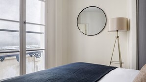 A hotel room with a large window, a round mirror, a floor lamp, and a bed with a dark blue blanket. - Blueground | Madeleine, elev, near art (Paris)
