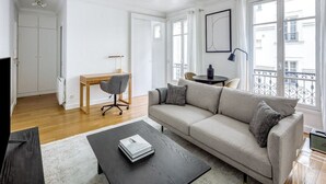 Un salon comprenant un canapé, une table basse et un bureau avec une chaise. Une fenêtre offre une vue sur les bâtiments.