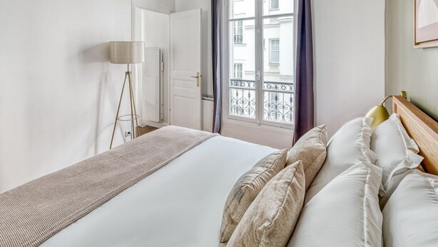 Une chambre d’hôtel avec un lit, une lampe sur pied, une fenêtre avec des rideaux et une vue sur les bâtiments environnants.