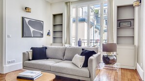 A living room with a grey sofa, a coffee table, and a clear side table with a lamp. There are shelves with books and a framed picture on the wall. - Blueground | Trocadero, concierge, nr Eiffel Tower (Paris)