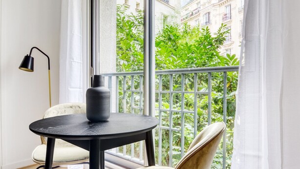 A room with a round black table, a black vase, a white chair, a floor lamp, and a balcony with a metal railing and greenery. - Blueground | Trocadero, intercom, nr Passy Plaza (Paris)