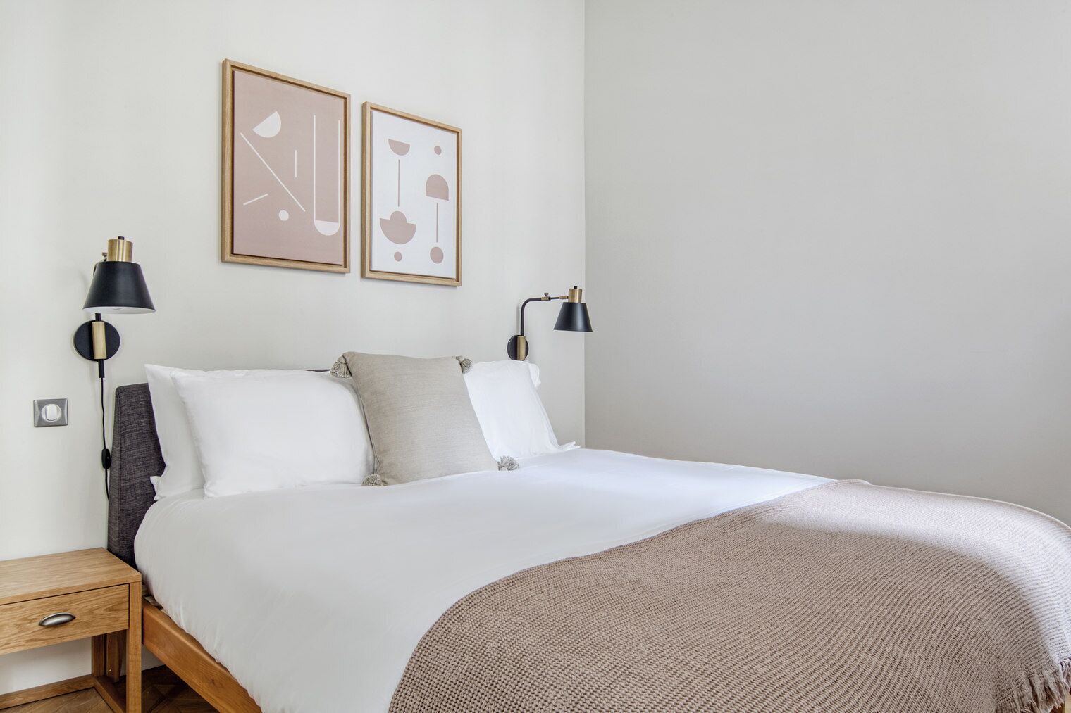 Una cama bien hecha con ropa de cama blanca y gris, dos lámparas fijadas a la pared y dos obras de arte abstractas enmarcadas.
