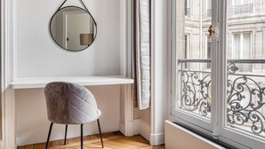 A room with a white desk, a round mirror, a grey chair, and a view of a balcony with wrought iron railings.