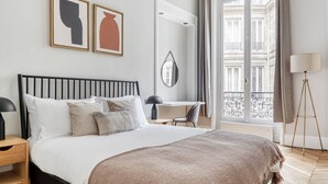 A modern hotel room with a bed, bedside table, desk, and a view of the city through a large window.