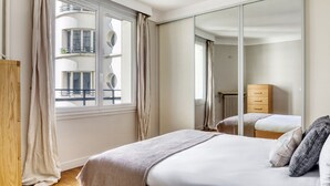 A bedroom with a large mirror, a bed with a grey blanket, a wooden dresser, and a window with curtains. - Blueground | Arc de Triomphe, furnished, elevator (Paris)