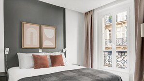 Ein Schlafzimmer mit einem großen Bett, zwei Kissen, einer grauen Decke, einem Fenster mit Blick auf einen Balkon und zwei gerahmten Bildern an der Wand.