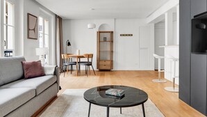 Ein modernes Wohnzimmer mit einem grauen Sofa, einem runden schwarzen Couchtisch und einem Essbereich mit Holztisch und Stühlen.