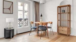 Ein modernes Zimmer mit einem runden Holztisch, vier Stühlen, einem schwarzen Beistelltisch mit Lampe, einem Holzregal und einem großen Fenster mit Vorhängen.