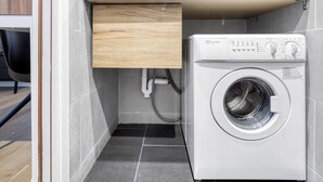 A compact laundry room with a front-loading washing machine, a wooden cabinet, and a visible pipe. - Blueground | Latin Quarter, nr Sorbonne (Paris)