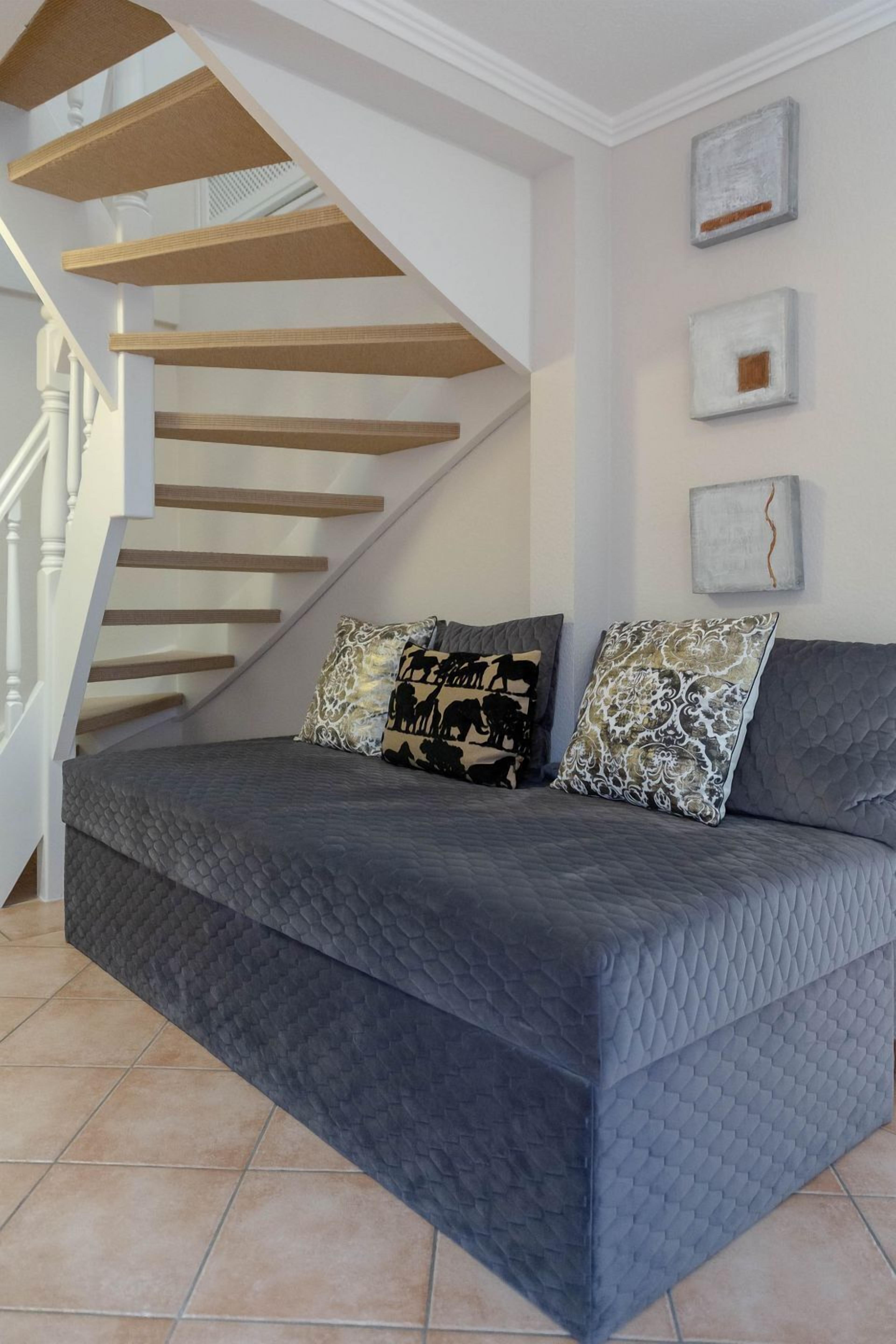 Un salon moderne avec un canapé gris à motifs matelassés, deux coussins à motifs, un escalier en bois et une rampe blanche.