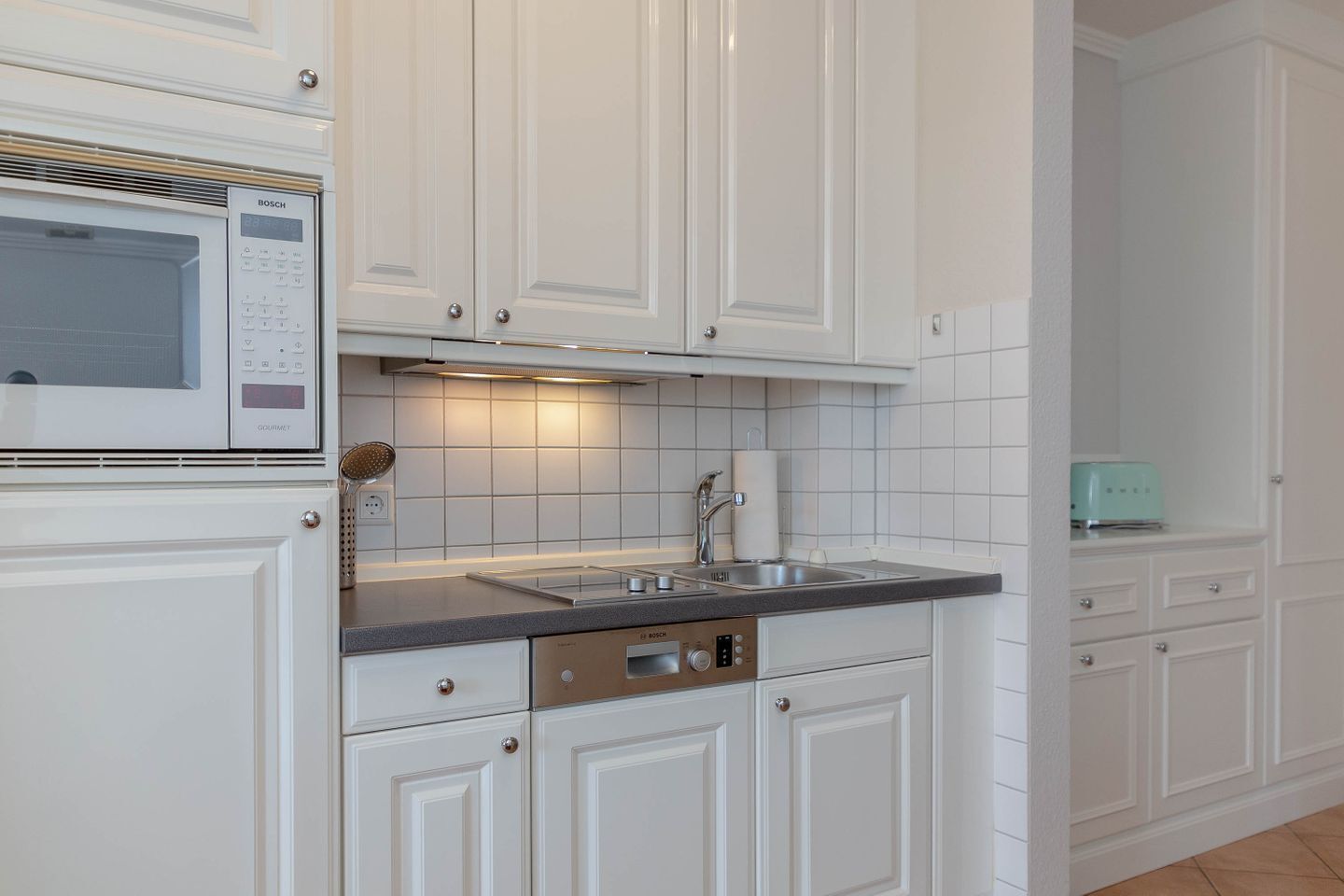 Una cucina con mobili bianchi, un microonde Bosch, forno integrato, lavello ed elettrodomestici inclusi lavastoviglie.