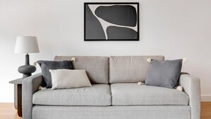 Un sofá gris con cuatro cojines, una mesita de noche con una lámpara y una obra de arte abstracta enmarcada en la pared.