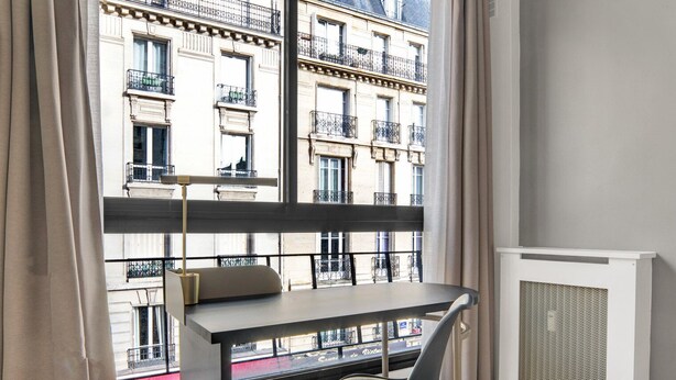 Une chambre moderne avec un bureau et une chaise, de grandes fenêtres donnant sur un immeuble doté de balcons.