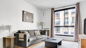 Un salon moderne avec un canapé gris, une petite table ronde, une table basse, une lampe et une grande fenêtre donnant sur des immeubles.