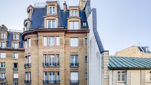 Un immeuble à plusieurs étages, de style architectural classique, caractérisé par une façade incurvée, de nombreuses fenêtres avec volets et un balcon d’angle bien visible.