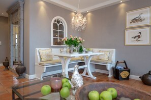 Un coin repas avec une table blanche, deux bancs, un lustre, des œuvres d’art encadrées et un vase rempli de fleurs.