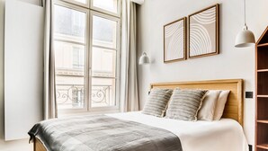 Un dormitorio con un ventanal grande, cabecera de madera, cama con manta estampada, dos almohadas y dos lámparas fijadas a la pared.