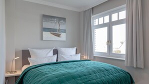 Ein Schlafzimmer mit Bett, Nachttisch, Lampe, einem Fenster mit Vorhängen und einem Bild an der Wand.
