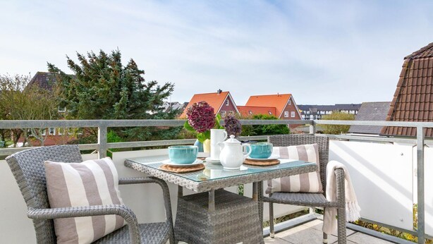 Ein Balkon mit Korbstühlen und einem Glastisch, mit Blick auf Häuser und Bäume im Hintergrund.