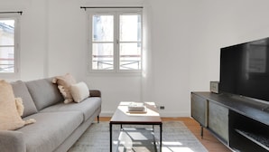 En moderne stue med en grå sofa, et sofabord, et fjernsyn på et skab og et vindue med persienner.