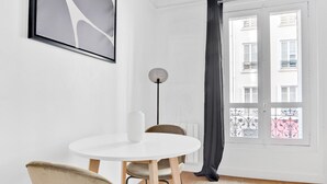 Una habitación minimalista con una mesa redonda blanca, una silla color beige, un jarrón blanco, una lámpara de pie y una ventana con vistas a edificios.