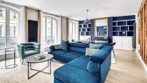 A modern living room with a blue sofa, a green armchair, and a round coffee table. There is a dining table with chairs, a bookshelf, and a large mirror on the wall. - Blueground | Rambuteau, elevator, nr Chatelet mall (Paris)